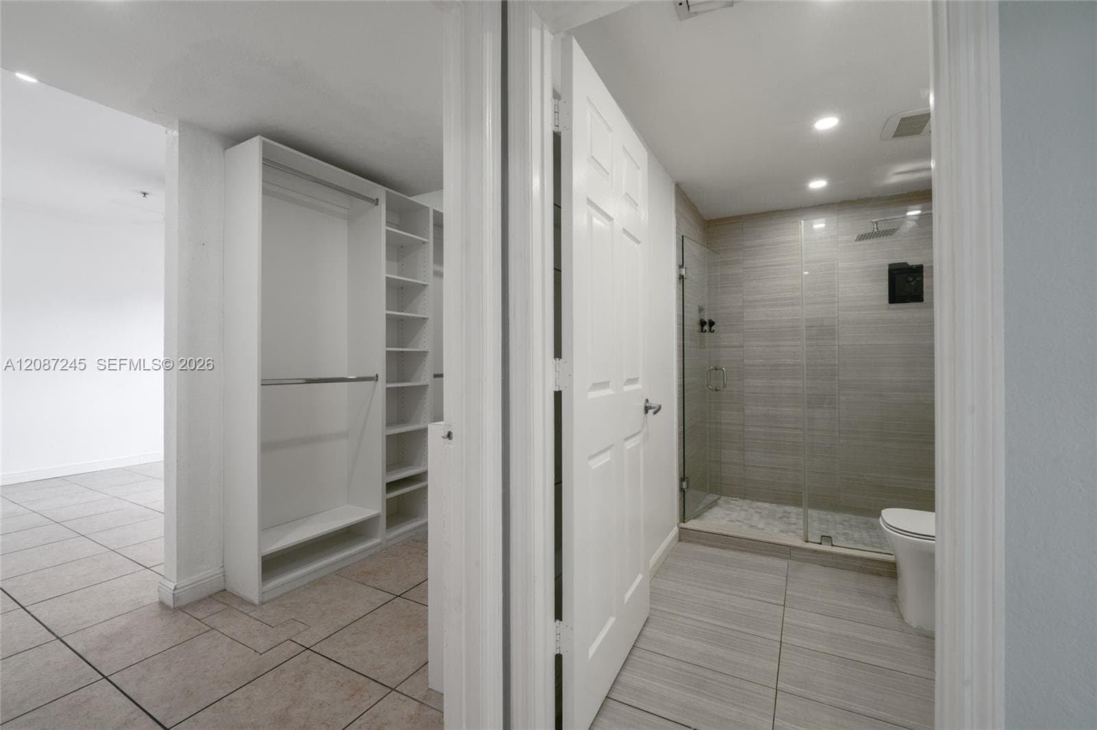 Walk-in closet featuring white built-in shelving and hanging rods, tiled flooring, glass-enclosed shower with horizontal wall tile, and recessed lighting