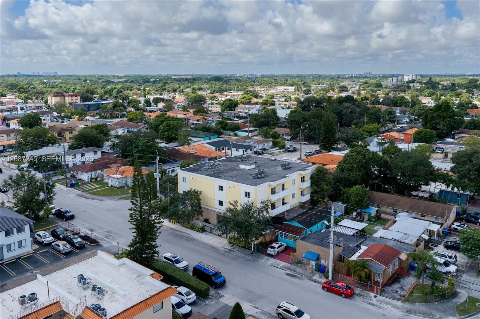 Aerial perspective of a multi-story residential building featuring a light-colored stucco exterior, flat roofline, and on-site surface parking, situated within an established urban neighborhood