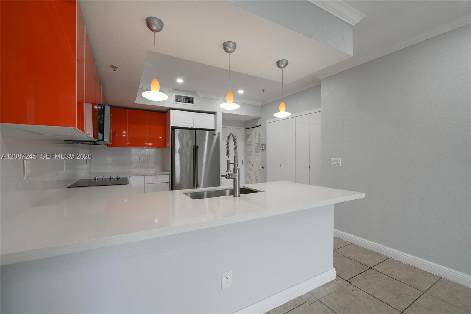 Modern kitchen featuring high-gloss orange cabinetry, quartz countertops, a stainless steel undermount sink with a gooseneck faucet, stainless steel refrigerator, and three glass pendant light fixtures