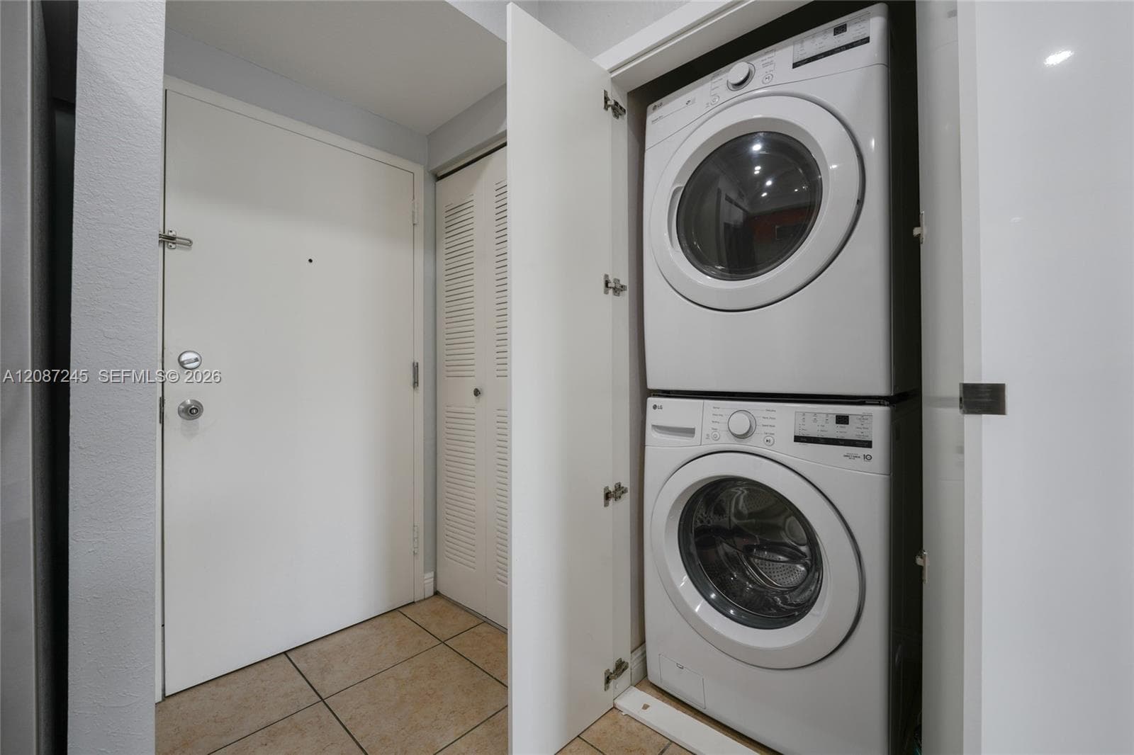 Stacked laundry unit set within a closet, featuring white louvered doors and tiled flooring