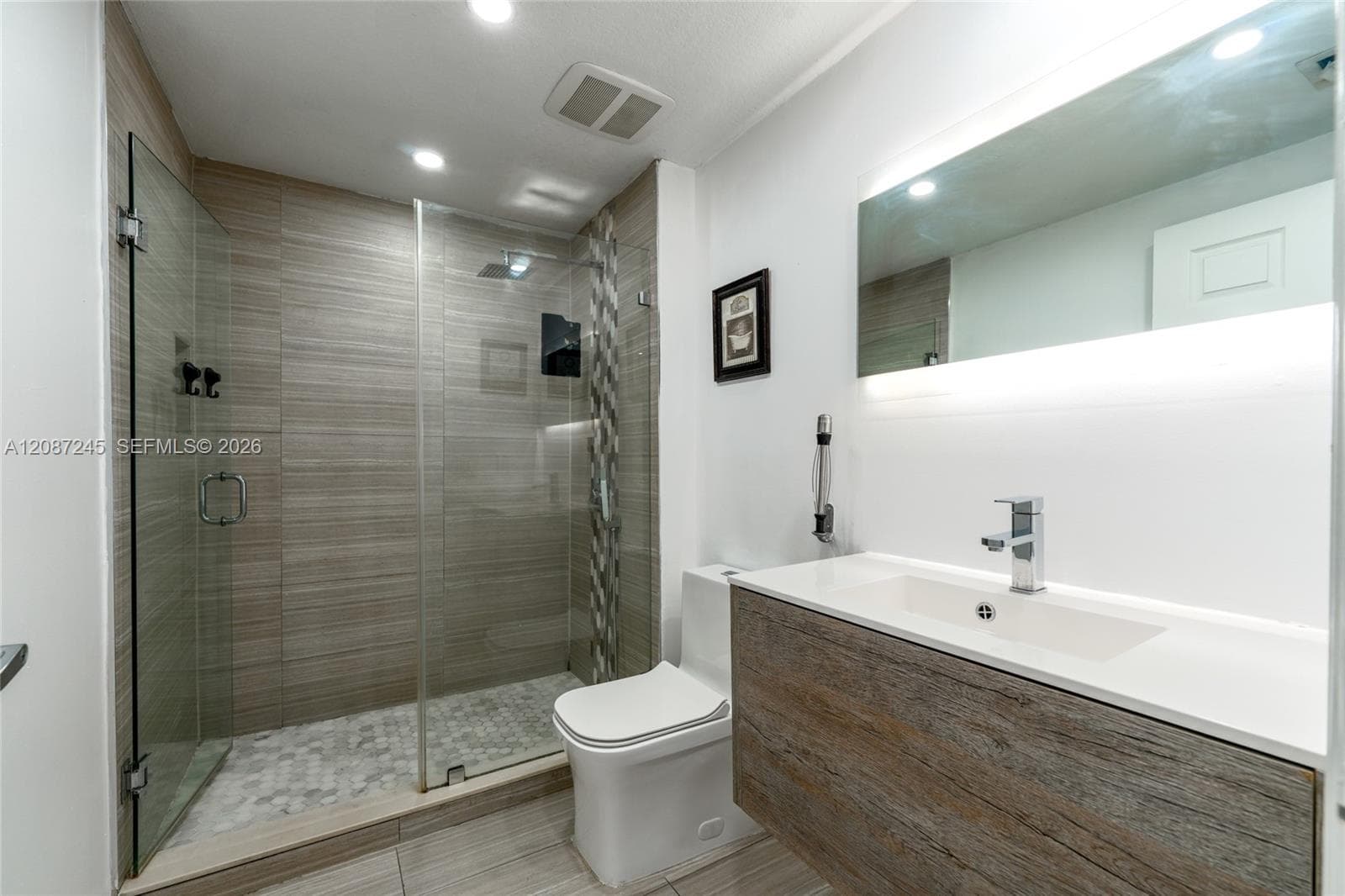 Floating wood-finish vanity with an integrated sink, frameless glass shower enclosure, horizontal stone-finish wall tiling, pebble-texture shower flooring, and backlit wall-mounted mirror
