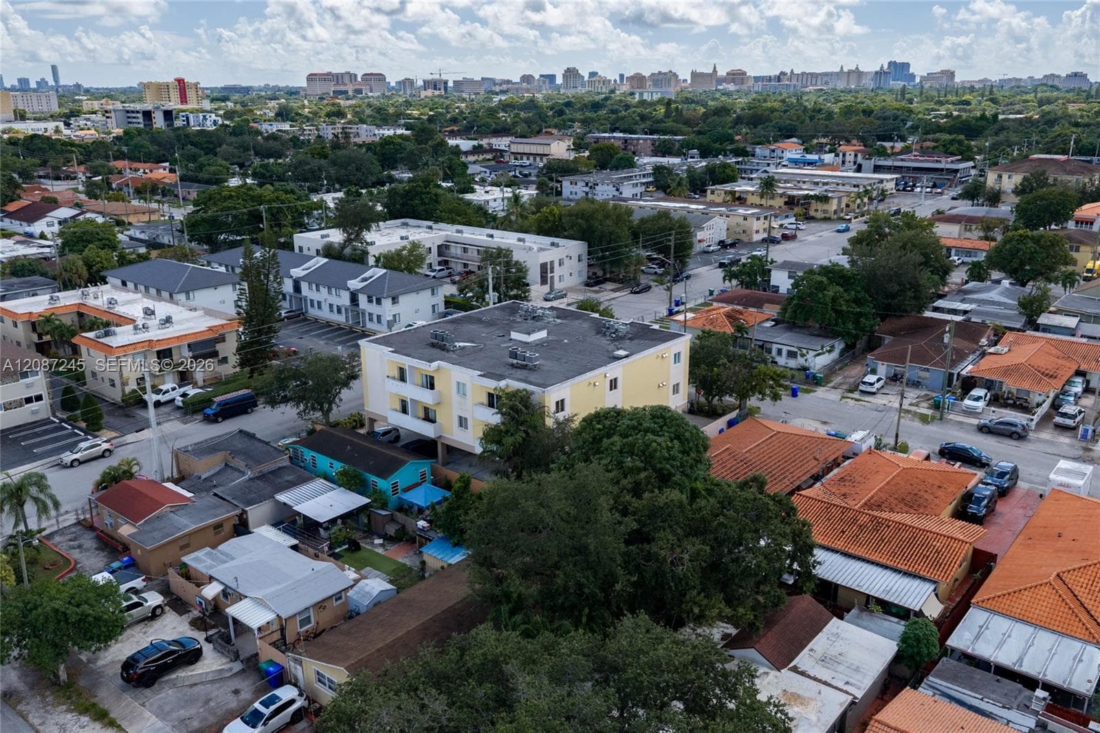 Aerial perspective of a multi-story residential building featuring a flat roof and balcony railings, situated in a dense urban environment with surrounding low-rise developments and mature trees