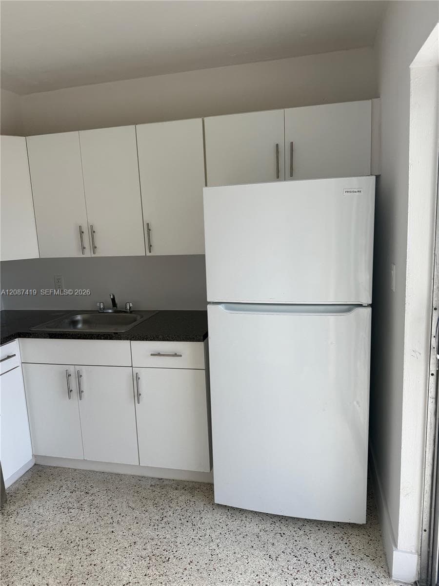 Kitchenette featuring white cabinetry with metallic pulls, dark stone-finish countertops, stainless steel sink with chrome faucet, top-freezer refrigerator, and terrazzo flooring