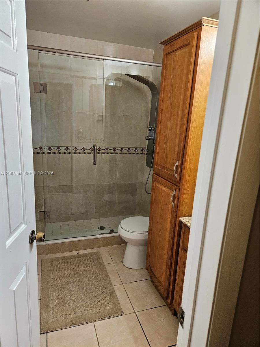 Walk-in shower with a glass enclosure, tile surround, and built-in shower panel system, accompanied by a tall wood-finish cabinetry unit and tile flooring