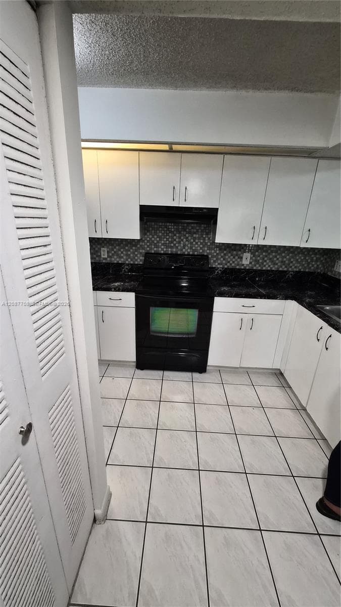 Kitchen featuring white cabinetry, dark countertops, patterned tile backsplash, black range, and light-toned tile flooring
