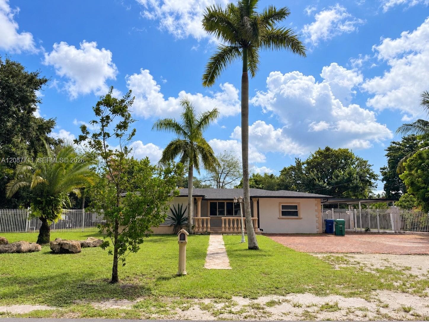 Single-story residence featuring a paved front walkway, decorative balustrade porch railing, exterior accent stone detailing, a brick-paved driveway, and mature palm landscaping