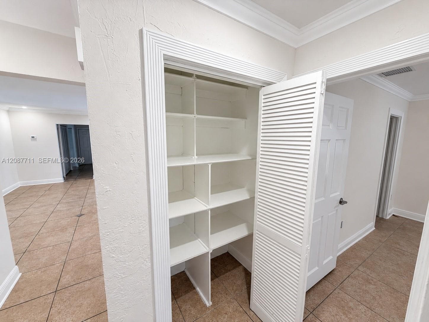 Built-in storage closet featuring custom shelving, louvered white door, decorative door casing, and tile flooring