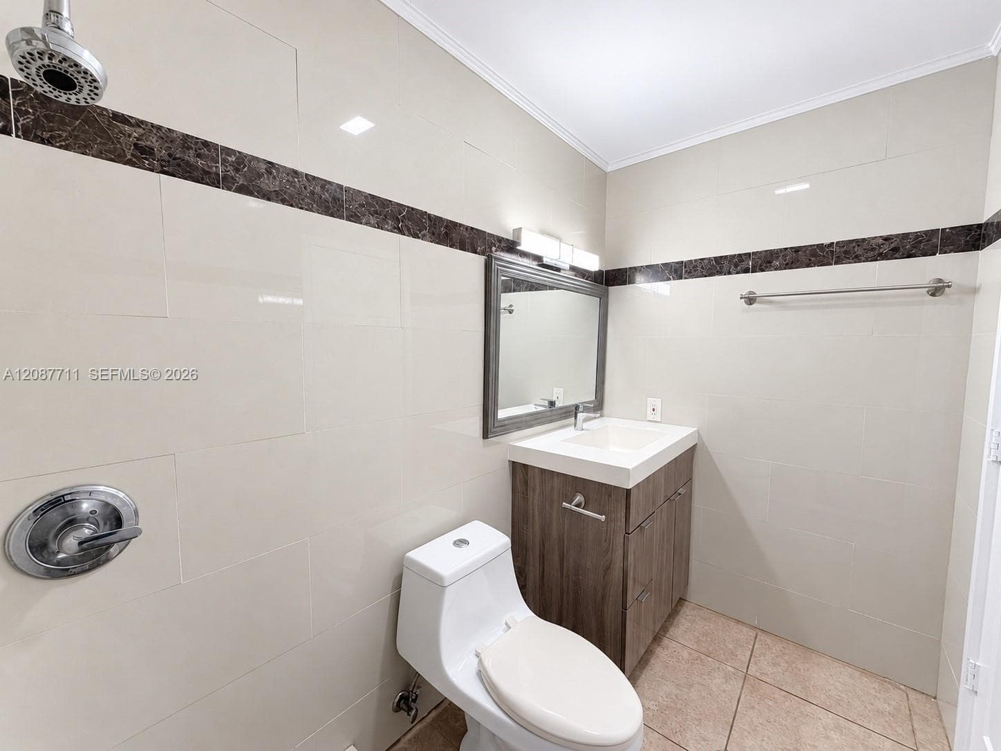 Bathroom featuring light-toned wall tile with dark stone accent trim, wood-finish vanity with a rectangular sink, wall-mounted light fixture, and metal towel bar