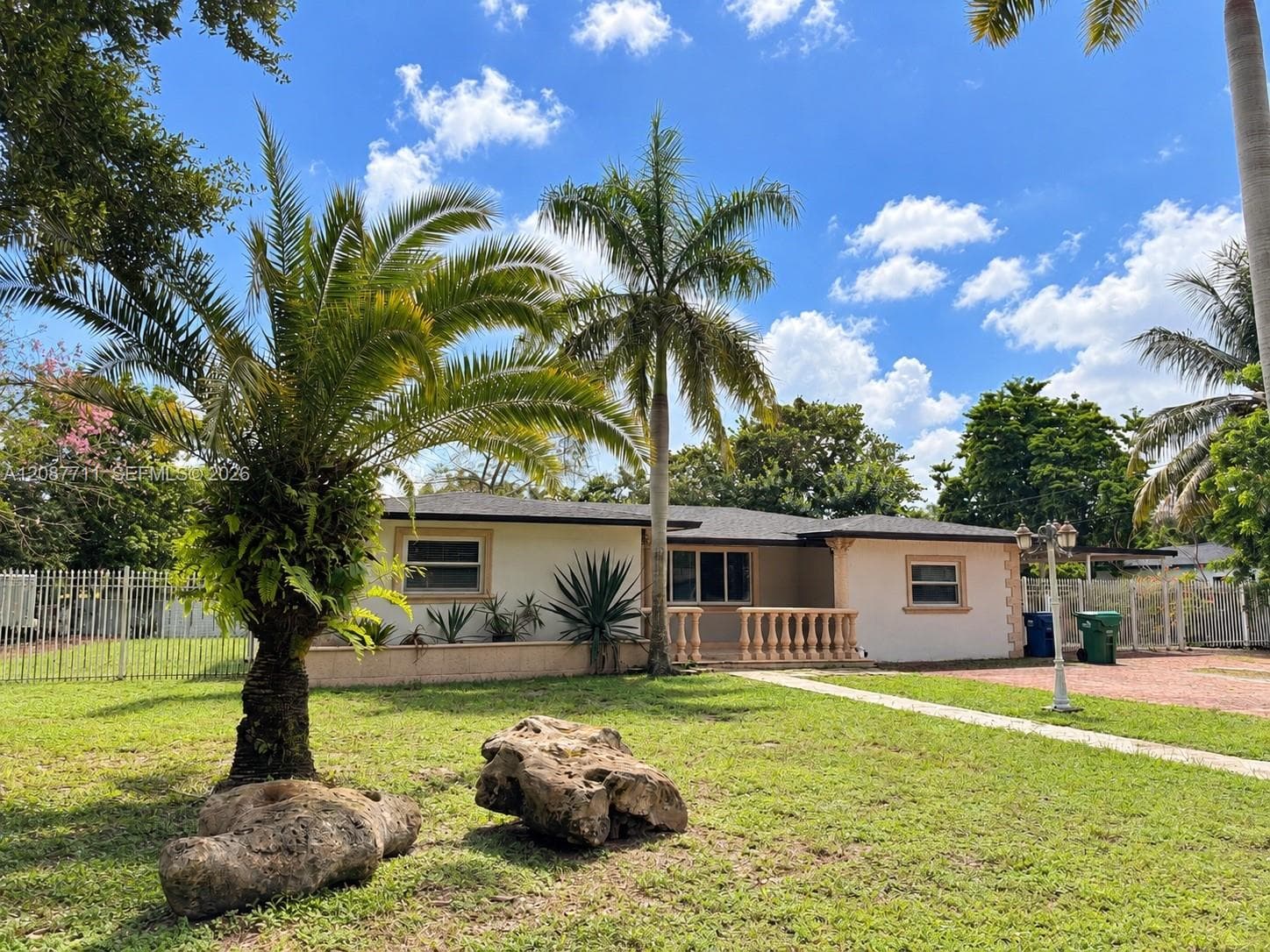 Single-story residence featuring a light-colored stucco exterior, decorative stone balustrade, mature palm trees, manicured lawn, and perimeter metal fencing