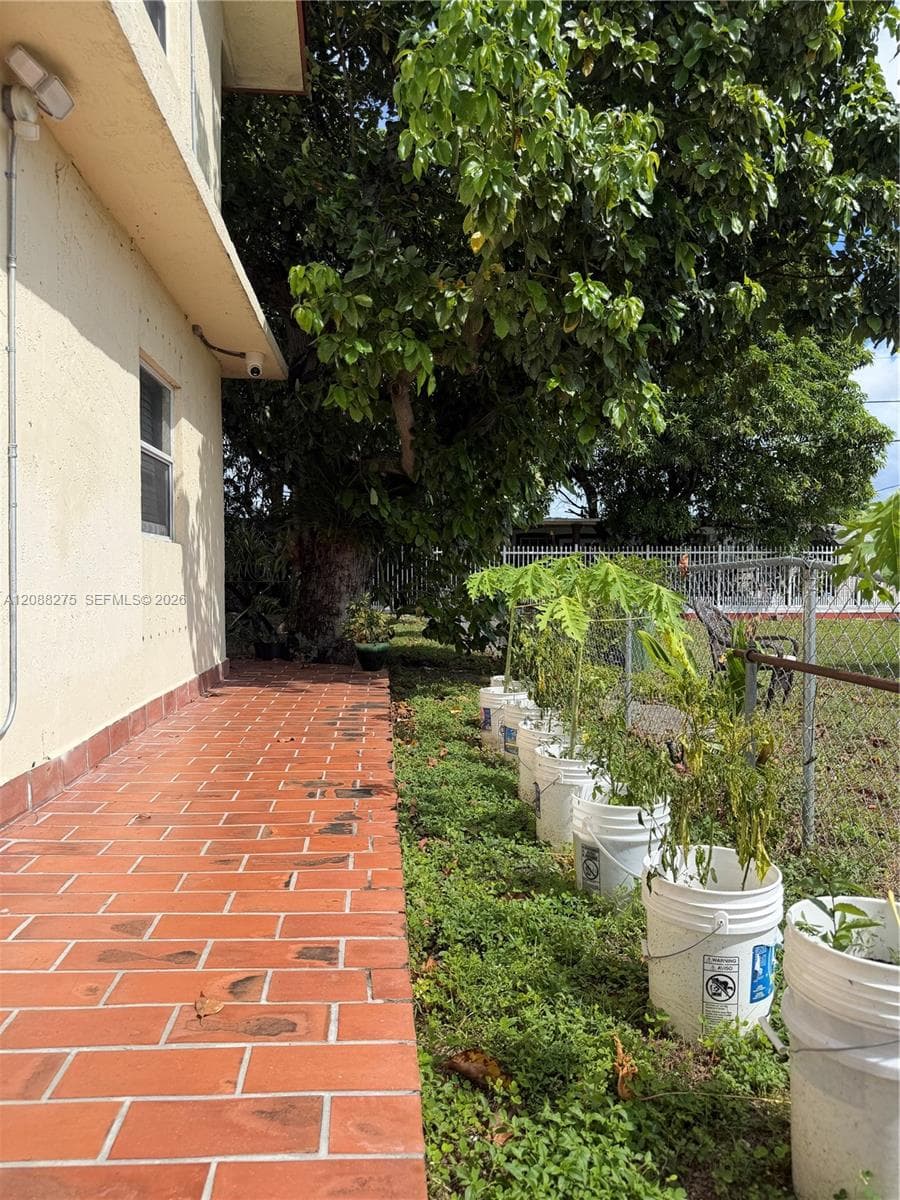 Exterior side yard featuring a terra cotta tiled walkway, stucco siding, a mature shade tree, and metal chain-link perimeter fencing