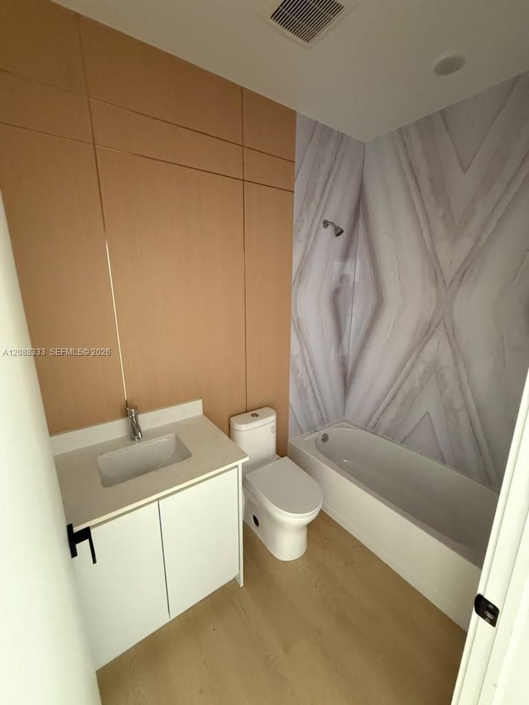Bathroom featuring a vanity with a white countertop and rectangular sink, wood-finish wall paneling, shower-tub combination with patterned surround, and wood-finish flooring