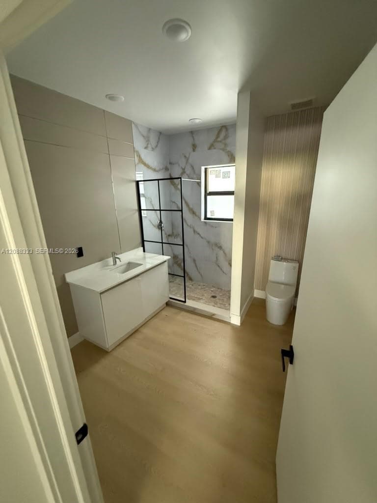 Bathroom featuring wood-finish flooring, white vanity with stone-composite countertop, and a walk-in shower with black-frame glass enclosure, patterned tile surround, and recessed lighting