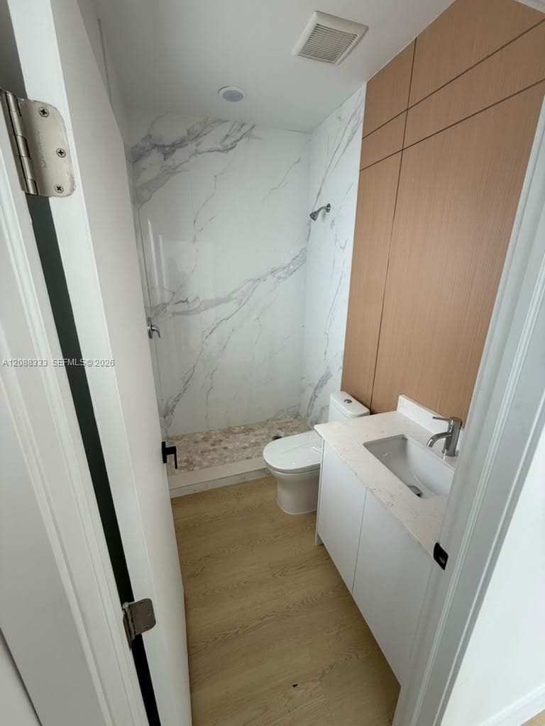 Bathroom featuring large-format wall panels with a white veined pattern, a pebble-finish shower floor, wood-finish flooring, a white vanity with a solid-surface countertop, and wood-finish vertical wall paneling