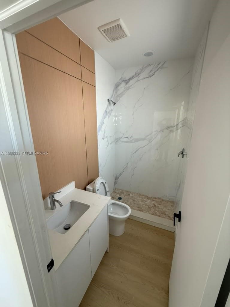Bathroom featuring a stone-finish shower surround, wood-finish wall paneling, wood-finish flooring, white vanity with a composite countertop, and a built-in shower head