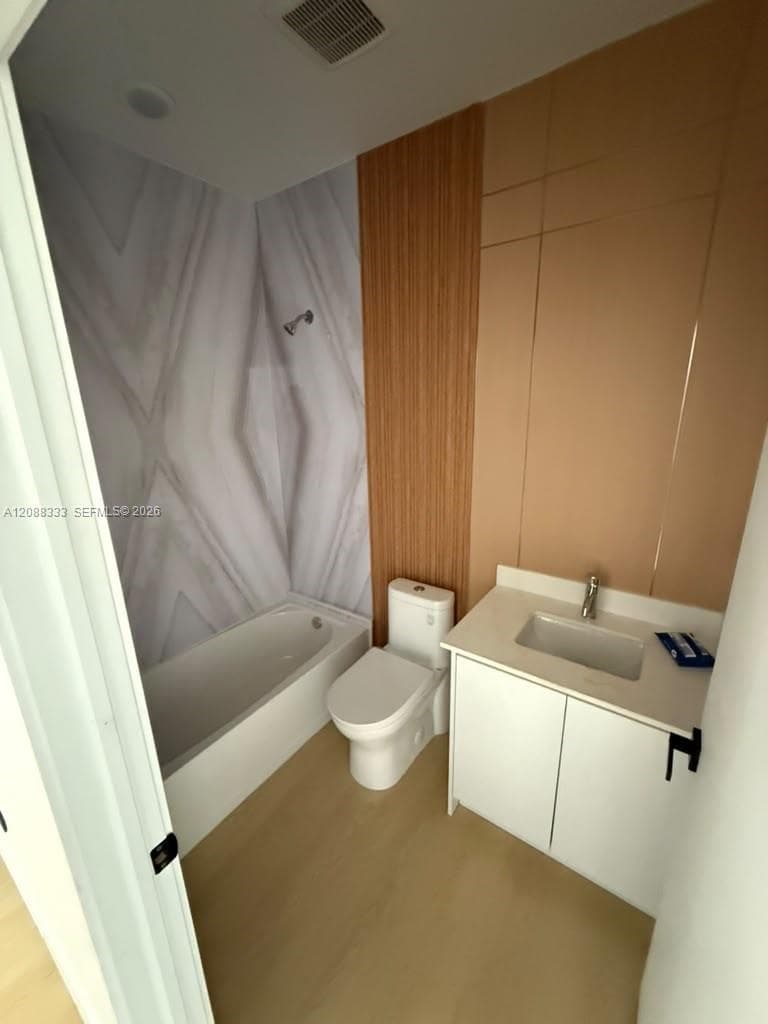 Bathroom featuring a built-in vanity with a stone-composite countertop, integrated sink, wood-finish wall paneling, and a tub-shower combination with patterned wall surround