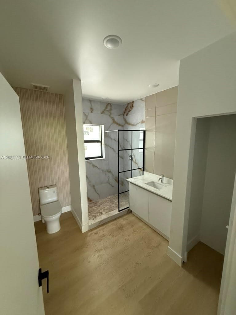 Bathroom featuring a walk-in shower with a black-framed glass enclosure, patterned tile surround, white vanity with a neutral countertop, and wood-finish flooring