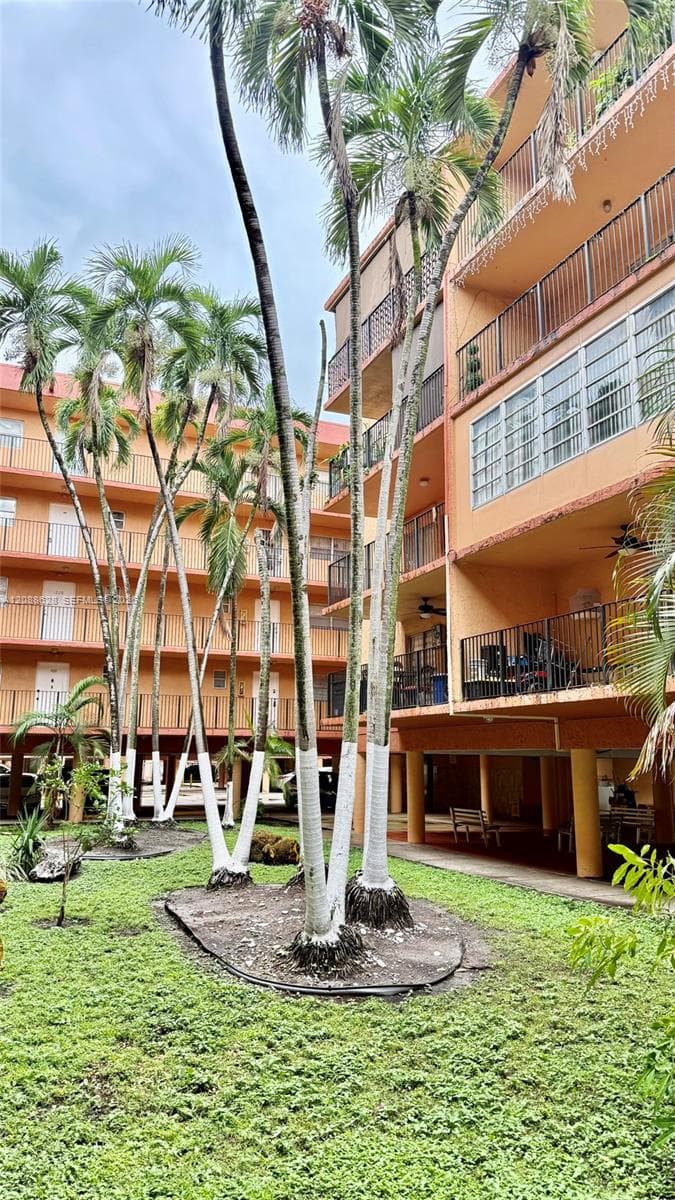 Mid-rise building featuring multi-level open-air balconies with metal railings, exterior staircases, and mature palm trees situated within a grass-covered common area