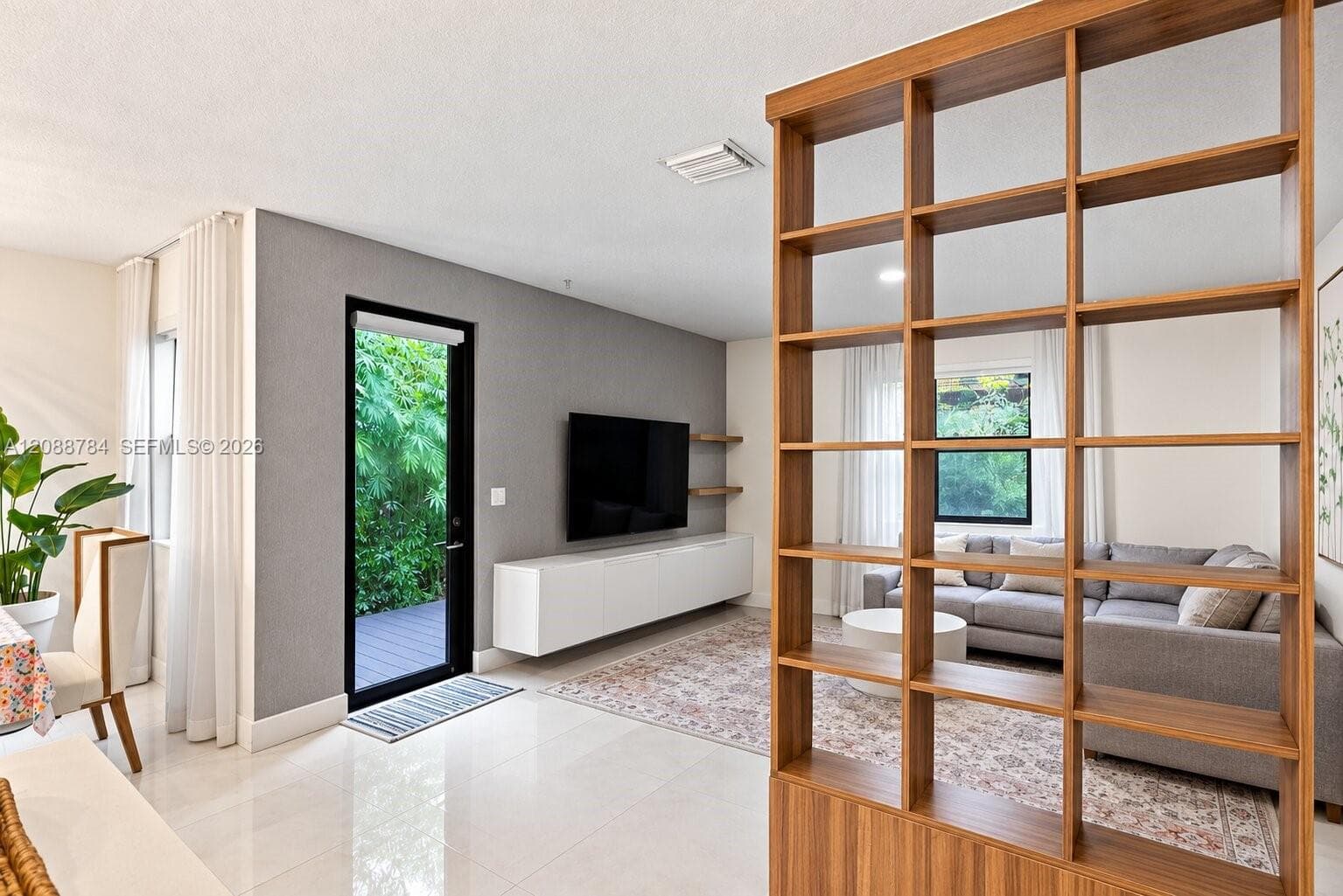 Polished tile flooring, a large window with privacy drapery, a built-in wood-finish shelving unit, a black-framed glass door, and a floating entertainment console
