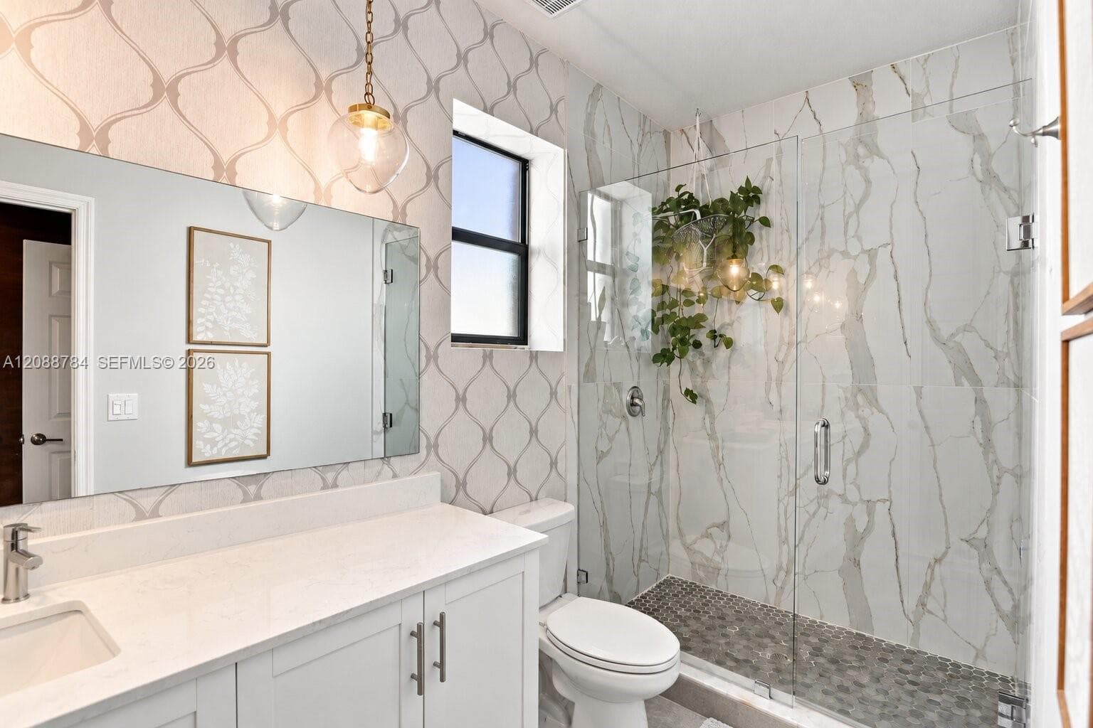 Bathroom featuring a stone-look tiled shower with a glass enclosure, pebble-tile shower floor, wall-to-wall vanity with a light-toned countertop, and patterned wall covering