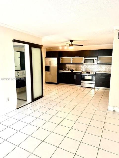 Kitchen featuring dark cabinetry, stainless steel appliances, glass-front upper cabinets, tile backsplash, and ceramic floor tiling