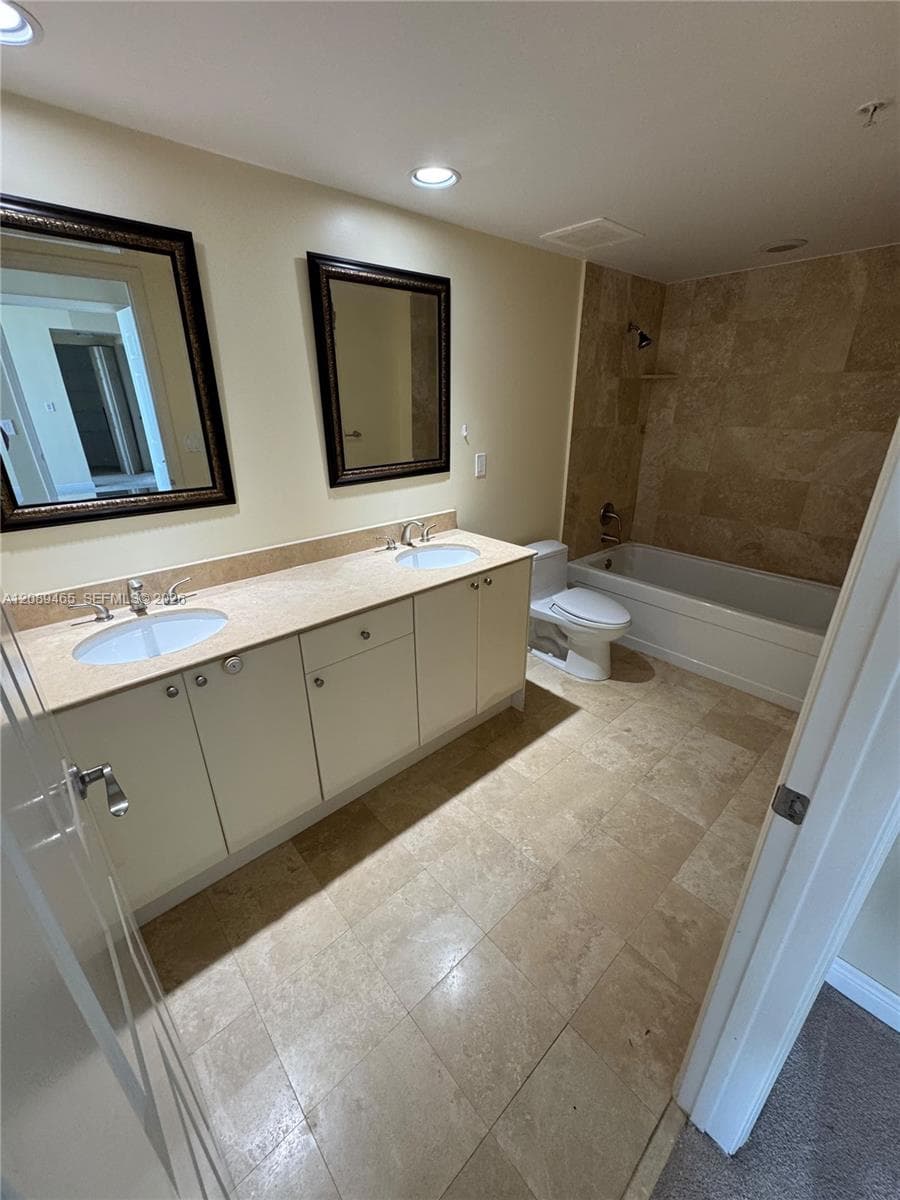 Dual-sink vanity with light-toned countertop, stone-tile flooring, and stone-tiled bathtub surround