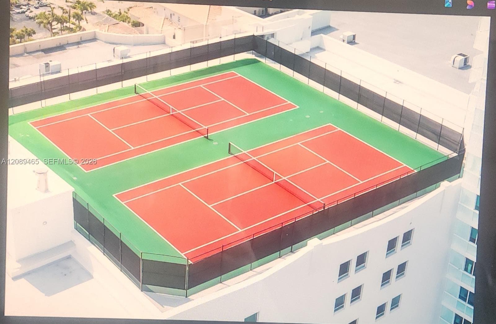 Rooftop amenity featuring two full-size tennis courts with red colored playing surfaces and green surrounds, enclosed by a perimeter fence