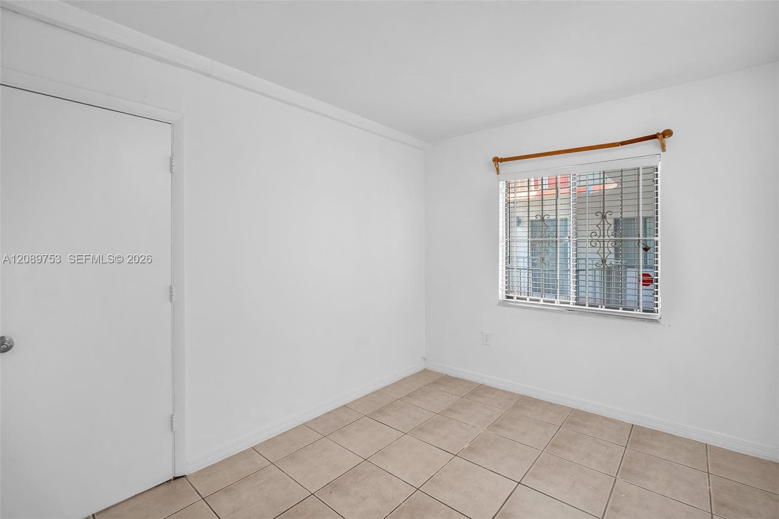 Neutral-toned ceramic tile flooring, solid-core interior door with metallic hardware, and a window featuring horizontal blinds and decorative security bars
