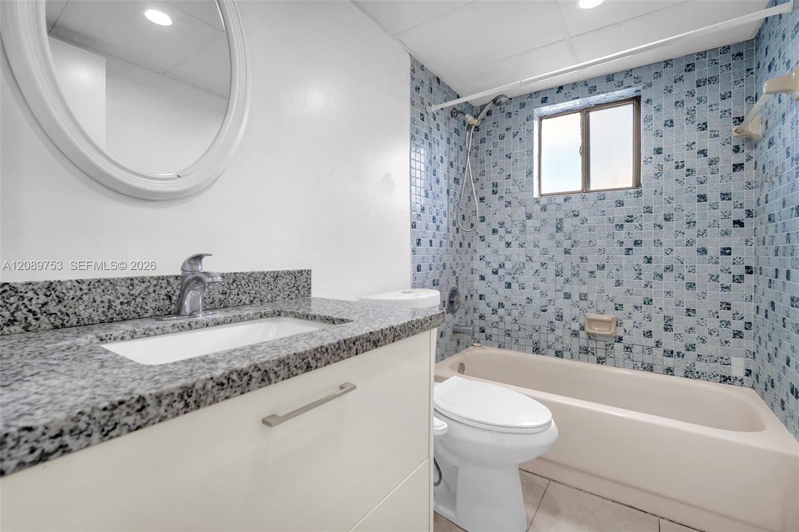 Bathroom featuring a vanity with a stone-finish countertop, undermount sink, oval wall-mounted mirror, and a shower-tub combination with blue mosaic tile surround