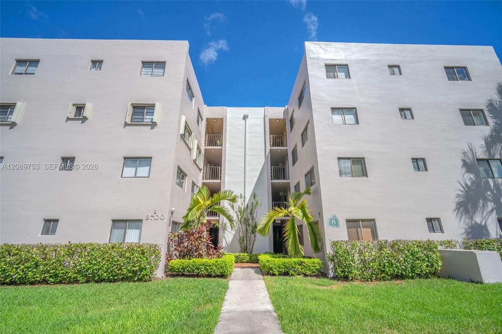 Multi-story exterior featuring a neutral stucco facade, inset balconies, manicured perimeter hedges, and a concrete pathway