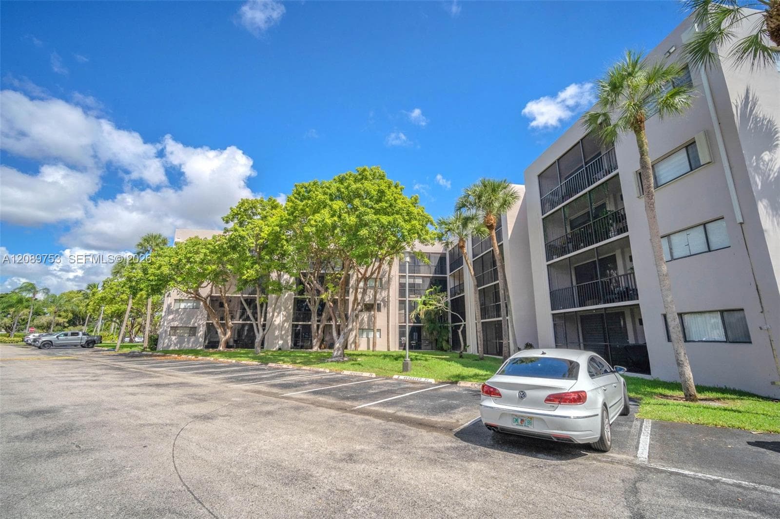Multi-story exterior featuring a light-toned stucco façade, private screened balconies, paved surface parking, and mature landscaping