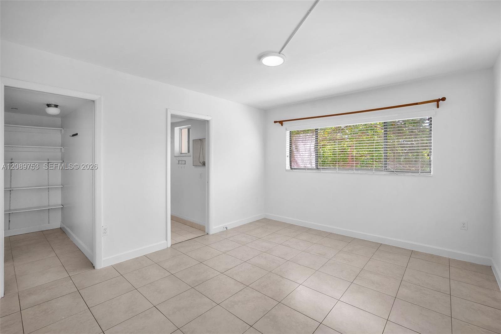 Neutral-toned tile flooring, large horizontal window with slatted blinds, and walk-in storage area featuring built-in shelving