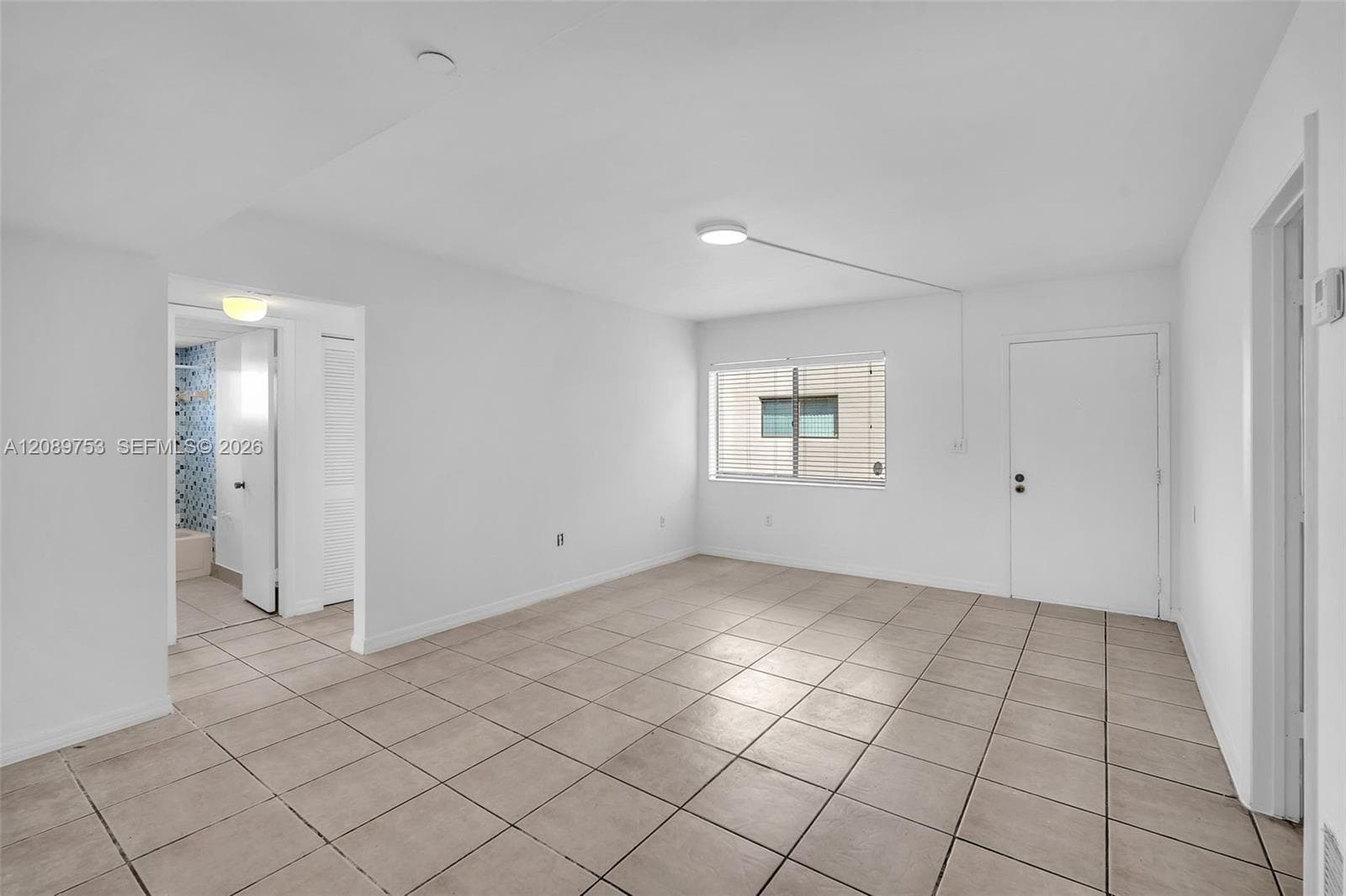 Neutral interior featuring large-format ceramic tile flooring, clean white walls, a single-pane window with horizontal blinds, and a semi-flush mount ceiling light fixture