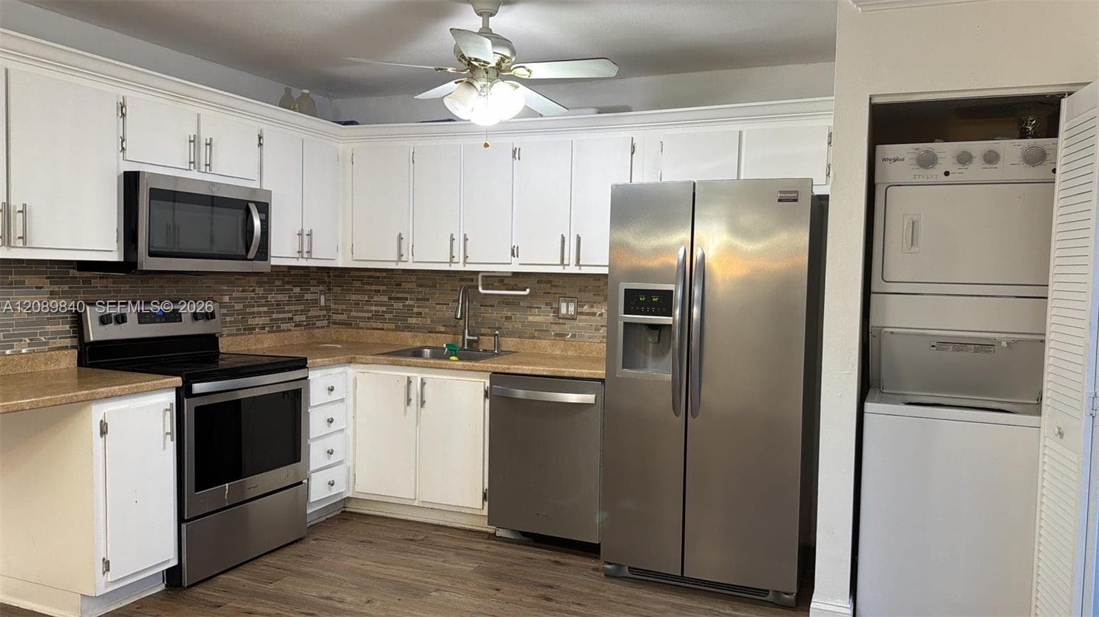 Kitchen featuring white cabinetry, stone-finish countertops, multi-tone tile backsplash, stainless steel appliances, and wood-finish flooring with a stacked laundry unit