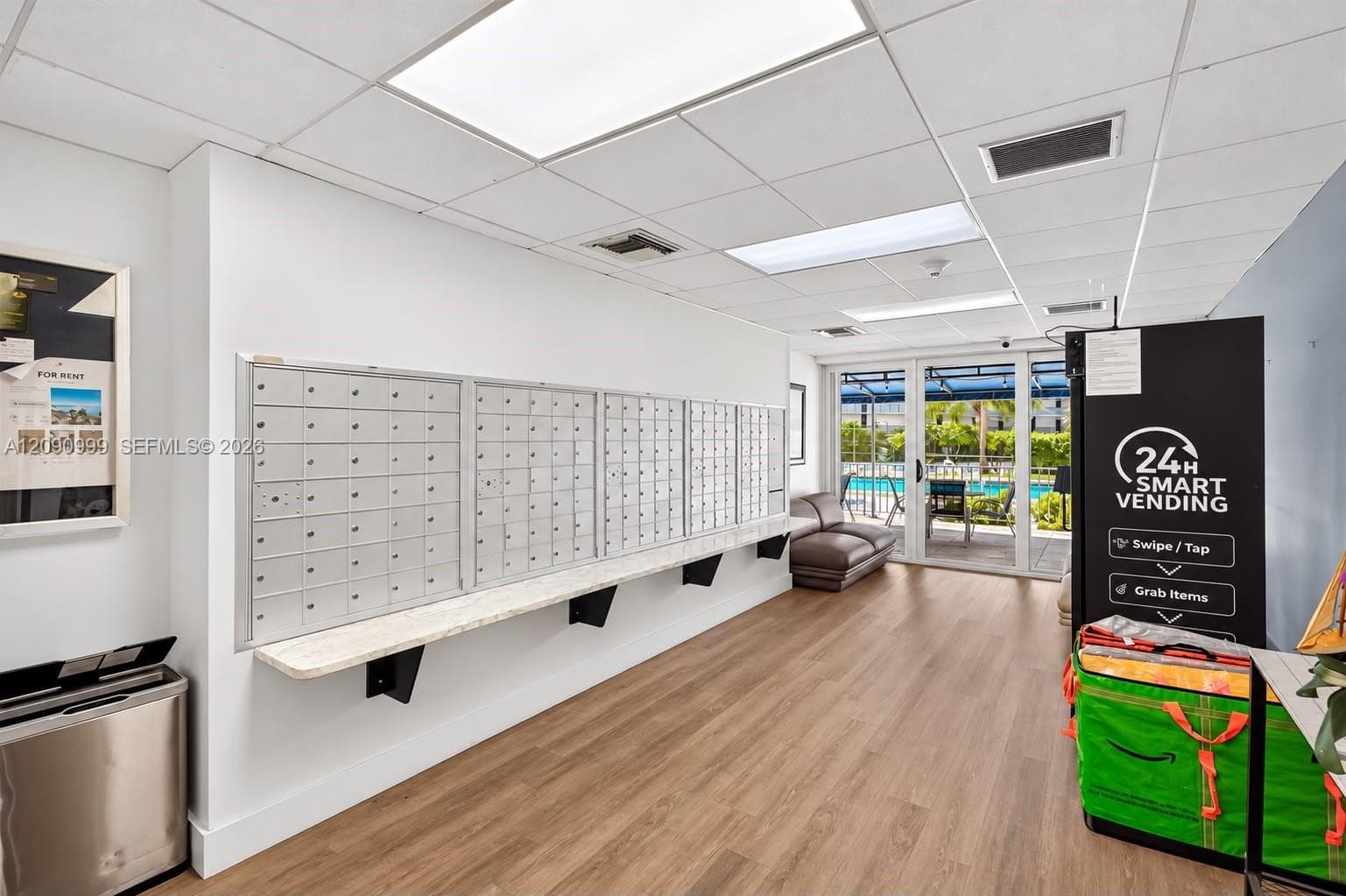 Wall-mounted mail collection center with integrated counter, wood-finish flooring, drop-ceiling panels with recessed lighting, and glass-paneled exterior door