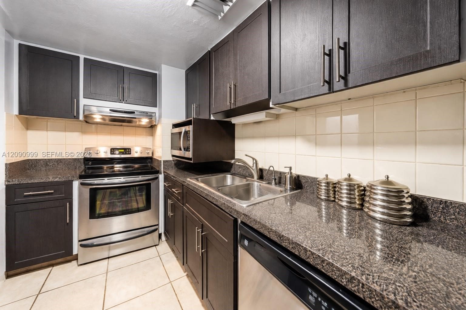 Kitchen featuring dark cabinetry, stone-surface countertops, stainless steel appliances, ceramic tile backsplash, and tile flooring