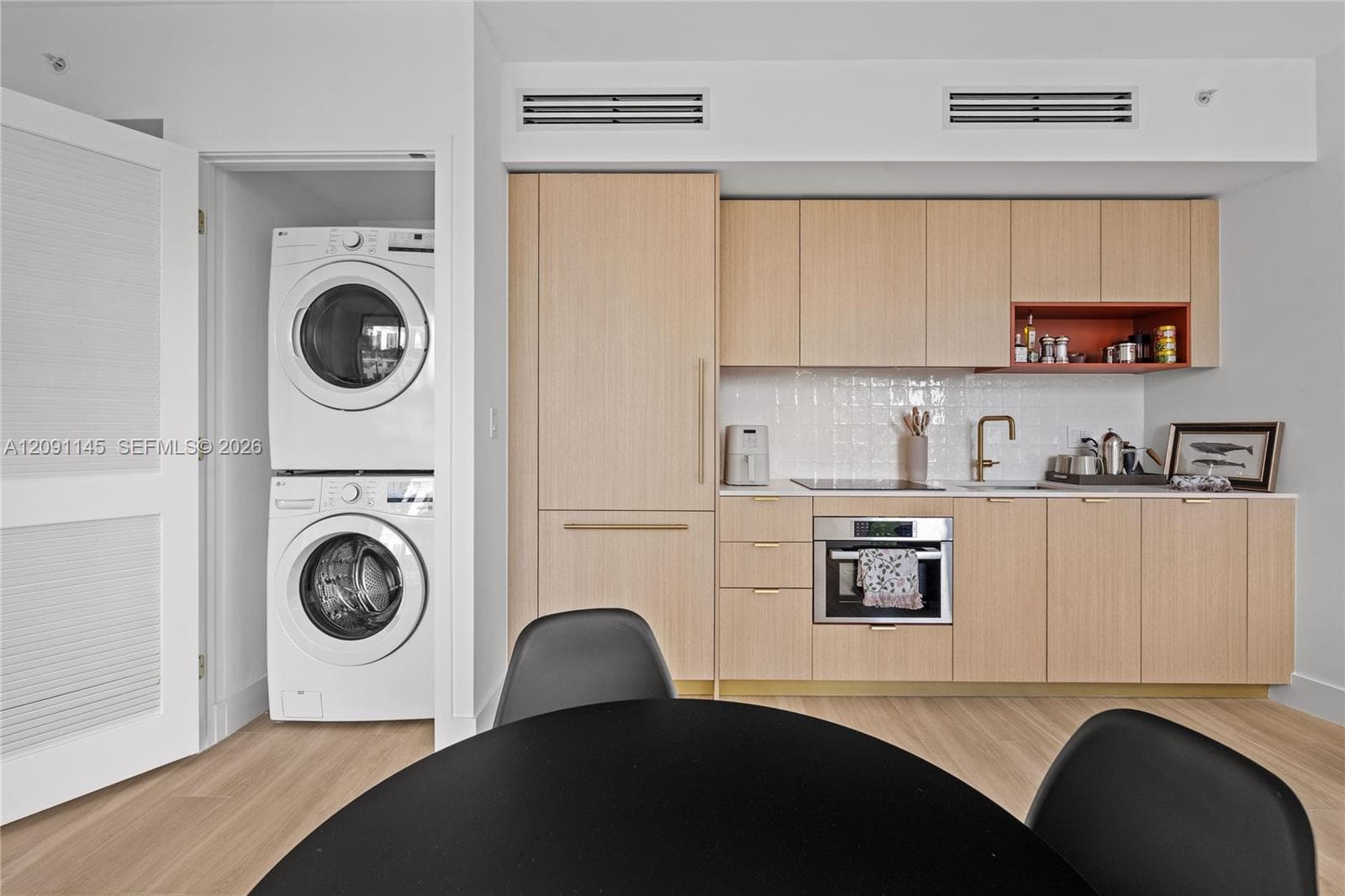 Integrated stackable washer and dryer, light wood-finish cabinetry, white tile backsplash, under-cabinet lighting, and stainless steel built-in oven