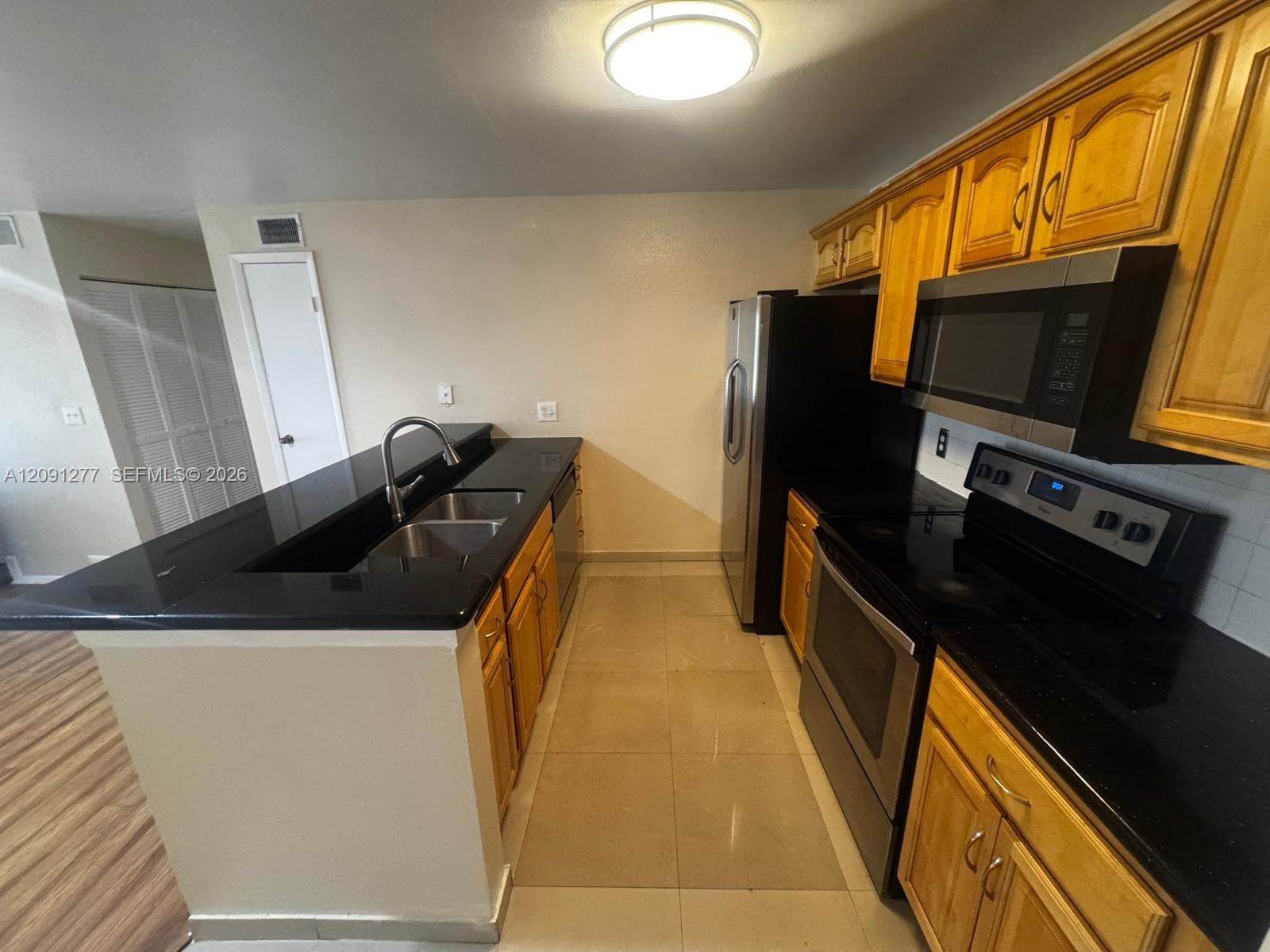Kitchen featuring wood cabinetry, black solid-surface countertops, stainless steel appliances, dual-basin sink with arched faucet, and ceramic tile flooring