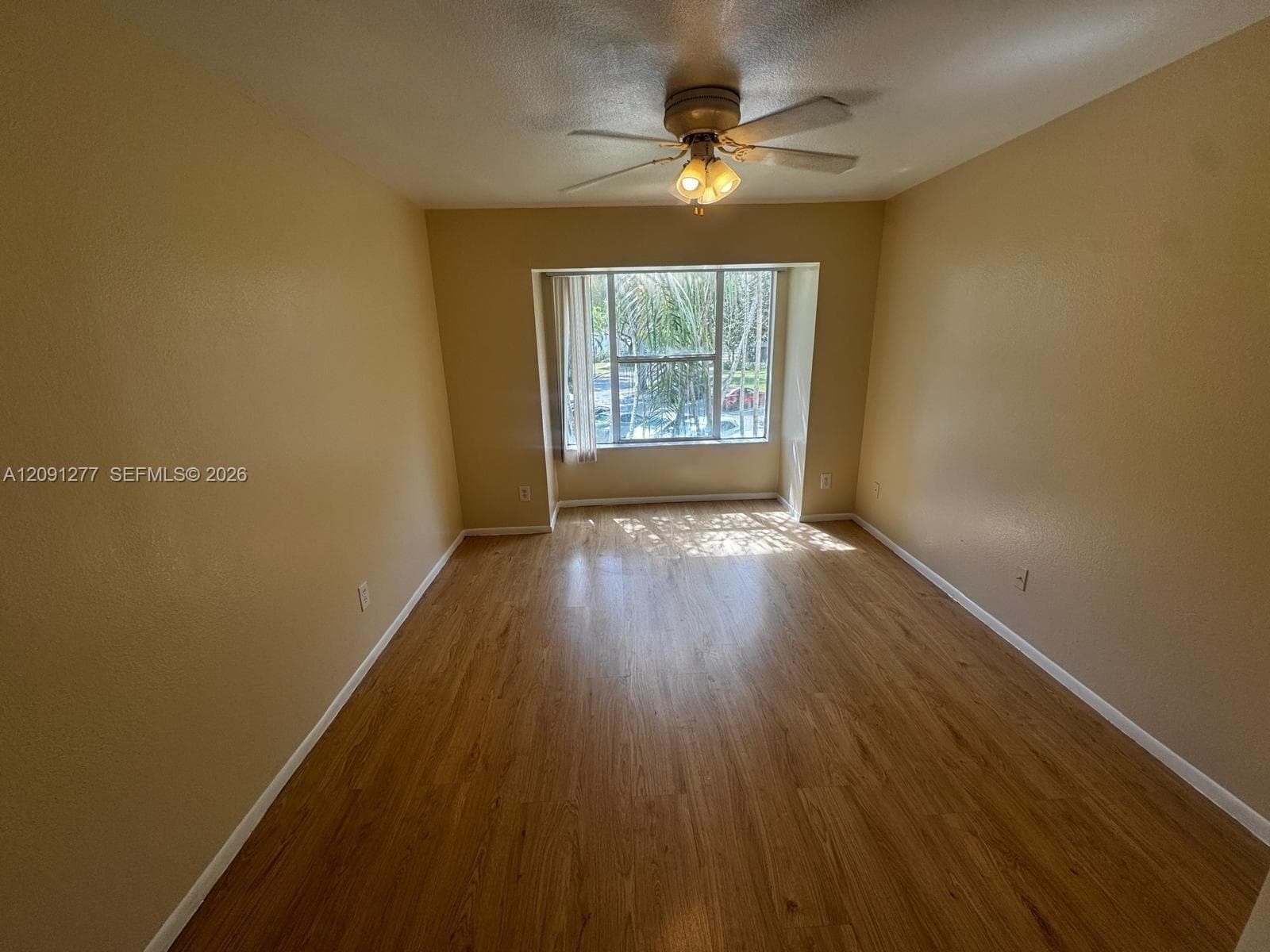 Neutral-toned interior space featuring wood-finish flooring, a large bay window, and a central ceiling fan with integrated lighting