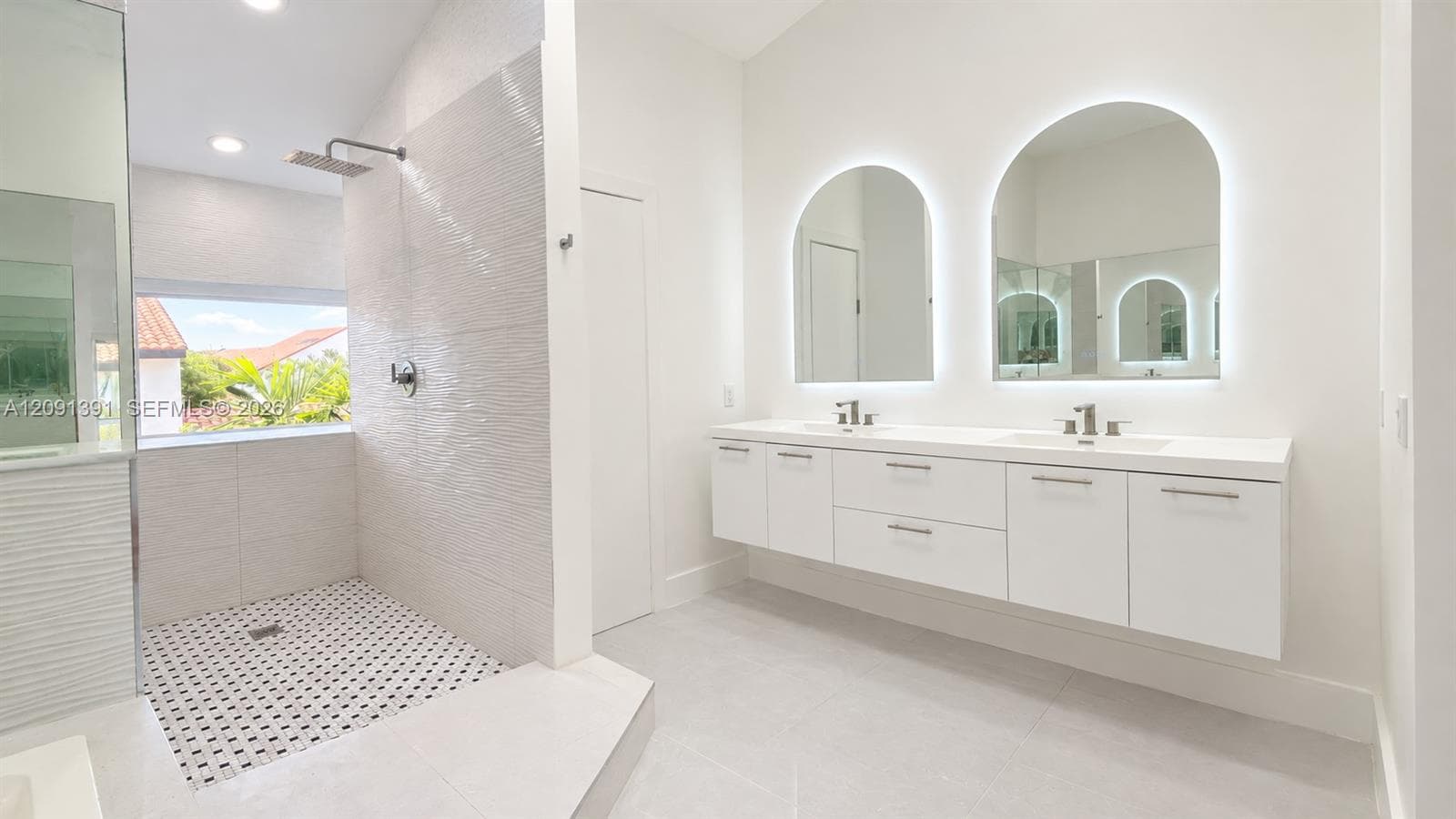 Floating double vanity with a clean-finish countertop, dual under-mount sinks, arched wall-mounted backlit mirrors, and a walk-in shower with textured tile detailing and a rainfall showerhead