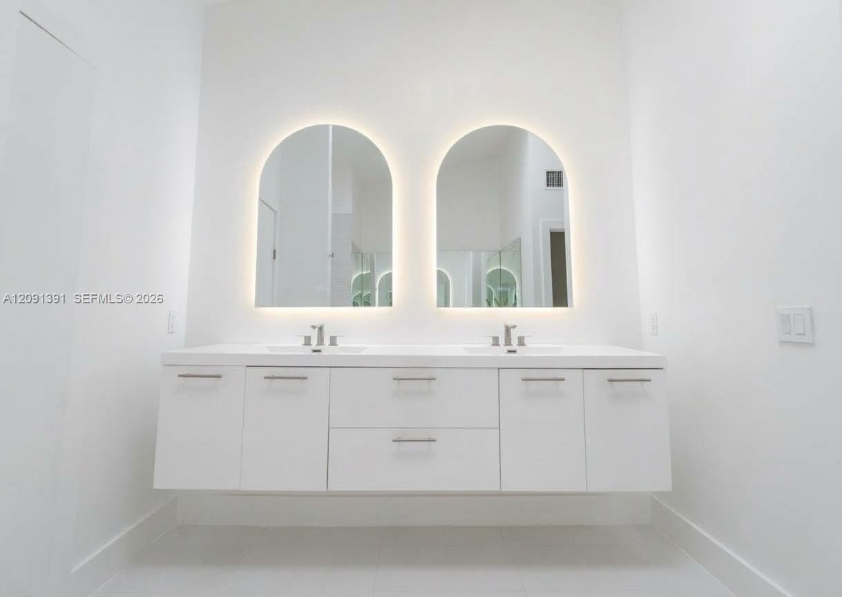 Floating double vanity featuring white cabinetry, dual integrated sinks, solid-surface countertop, and arched LED-backlit mirrors with polished chrome fixtures
