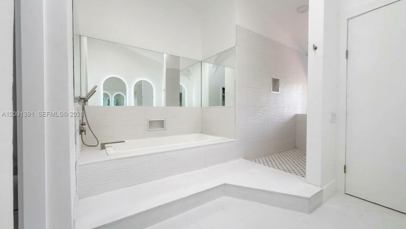 Platform-mounted soaking tub with an integrated hand-held shower fixture, large-format mirror wall, custom built-in recessed niche, and walk-in shower featuring geometric tile flooring