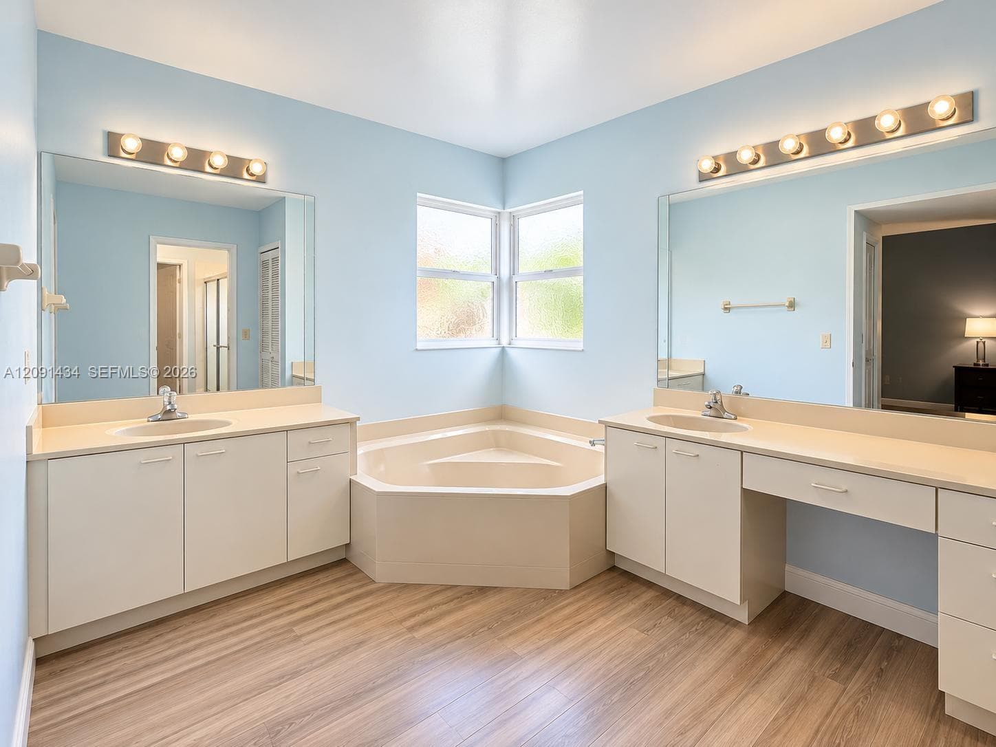 Dual vanity area featuring light-toned cabinetry, solid-surface countertops, corner soaking tub, large wall-mounted mirrors, and wood-finish flooring