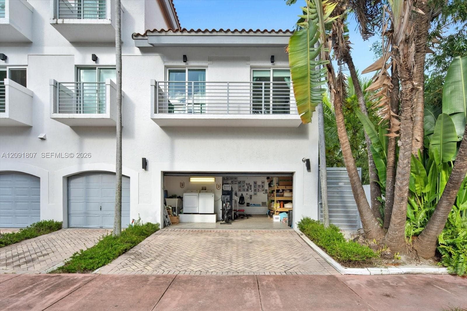 Stucco exterior featuring dual-level metal balconies, terracotta-style roof tile accents, a private herringbone-patterned paver driveway, and an attached multi-car garage
