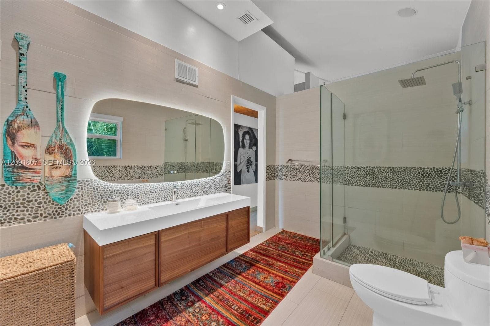 Floating wood-finish vanity with a white countertop and integrated sink, illuminated irregular-shaped vanity mirror, glass-enclosed shower with pebble tile accents, and large-format neutral wall and floor tiling