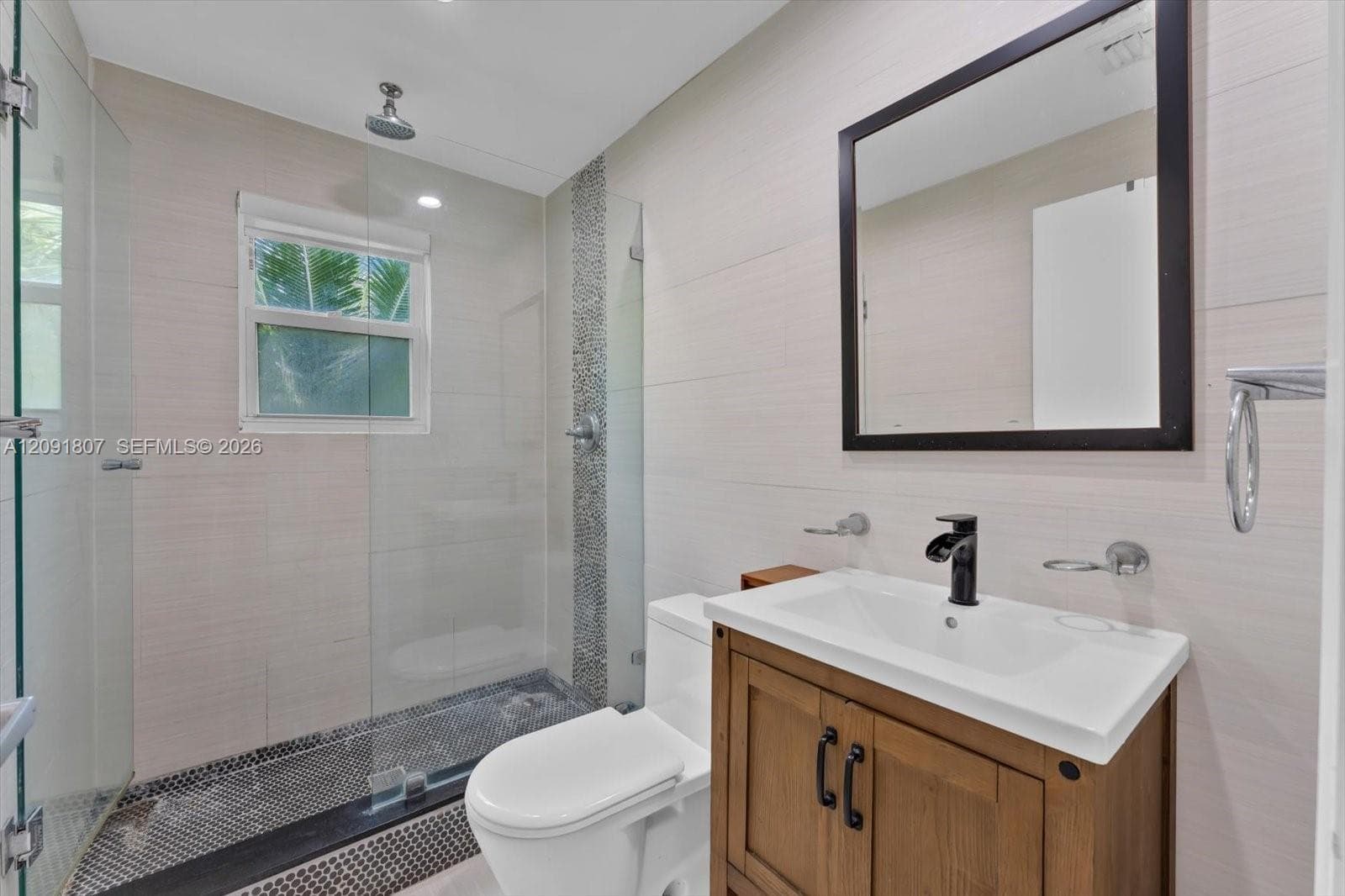 Bathroom featuring a wood-finish vanity with an integrated sink, matte black faucet, walk-in shower with a glass enclosure, pebble-tile flooring, and large-format wall tiling