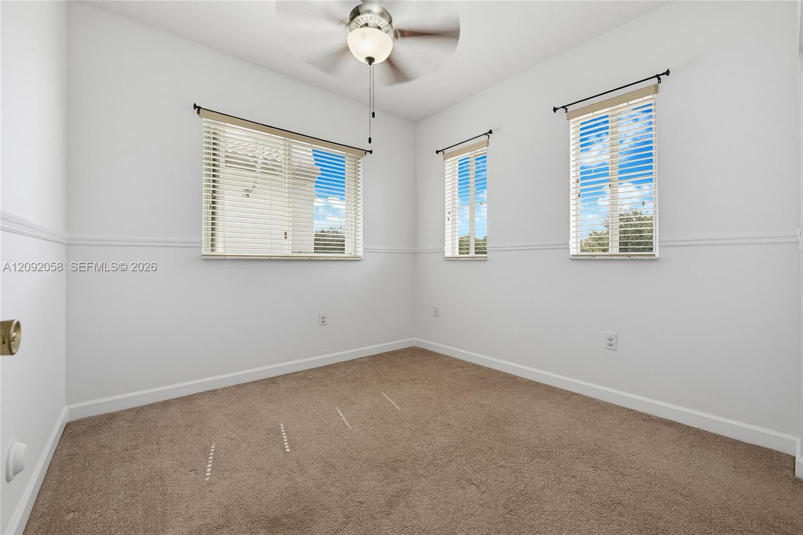 Neutral wall-to-wall carpeting, chair rail molding, three windows with horizontal blinds, and a ceiling-mounted fan with integrated lighting