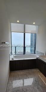 Soaking tub featuring a deep basin, dark cabinetry, stone-finish flooring, recessed lighting, and floor-to-ceiling windows