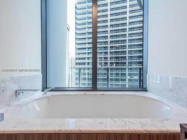 Built-in soaking tub with a stone-surround deck, polished metal wall-mounted faucet, and floor-to-ceiling windows featuring a high-rise exterior backdrop