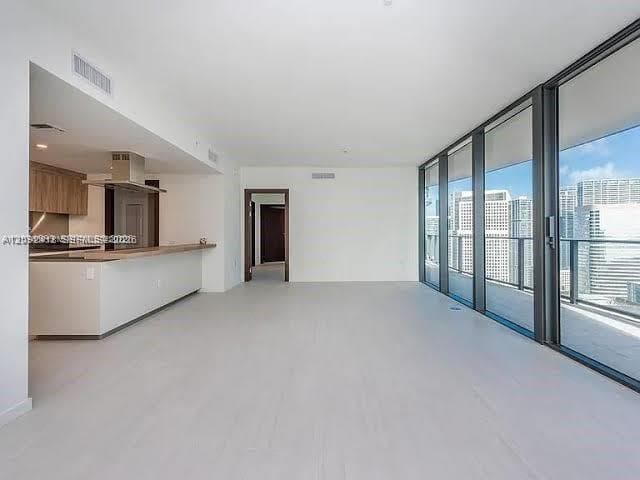 Open-concept interior featuring floor-to-ceiling glass panel walls, light-toned tile flooring, kitchen with wood-finish cabinetry, stainless steel range hood, and breakfast bar