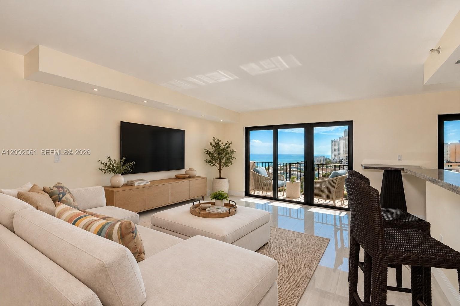 Open-concept living space featuring high-gloss tile flooring, recessed ceiling lighting, a built-in stone-top breakfast bar, and expansive multi-panel sliding glass doors opening to a balcony with an ocean backdrop